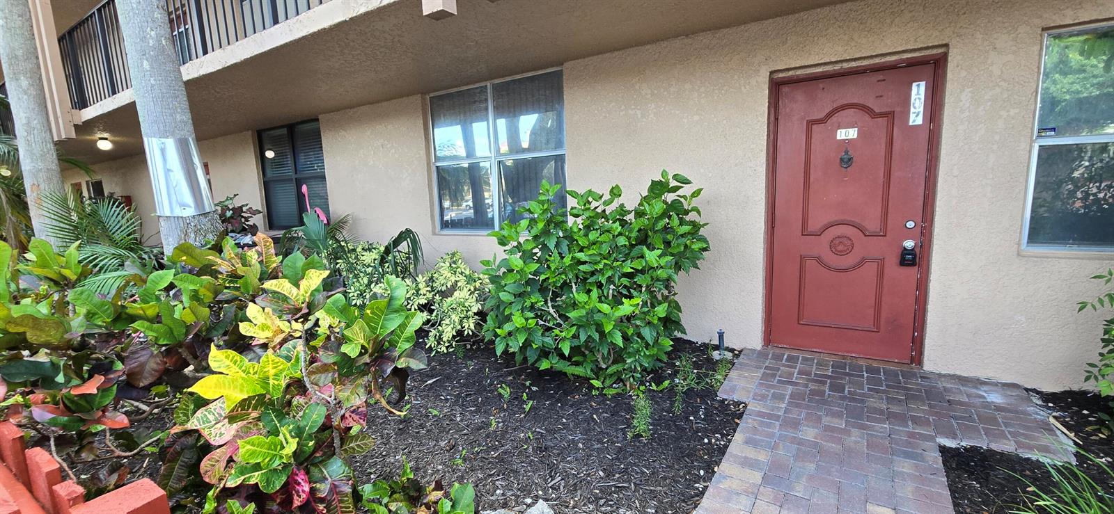 An exterior view of a ground-floor apartment entrance with a red door and landscaping — 7300 Lake Cir Drive # 107, Margate FL 33063, MARGATE, FL