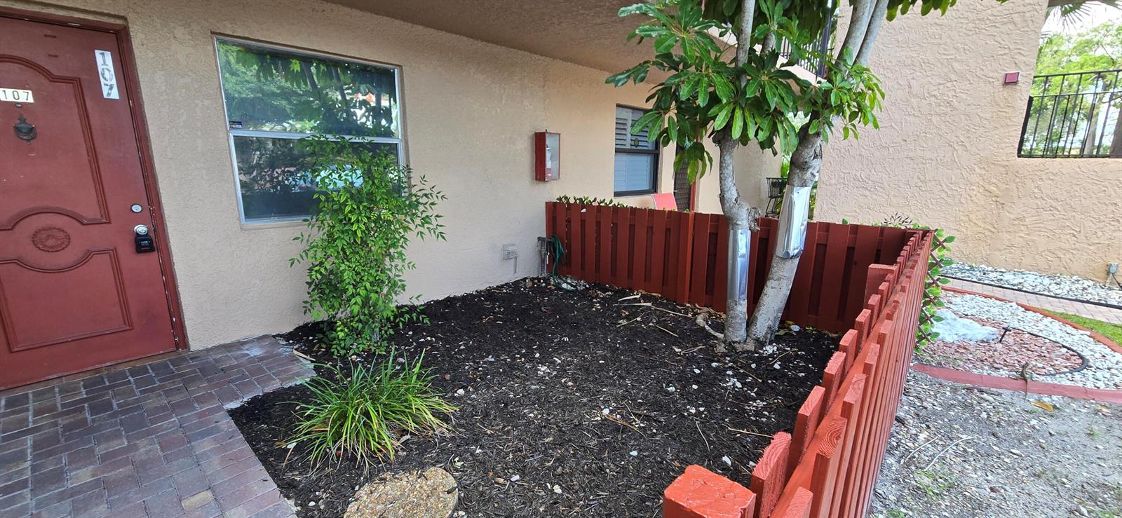 A small garden area with mulch, a tree, and a red fence outside an apartment — 7300 Lake Cir Drive # 107, Margate FL 33063, MARGATE, FL