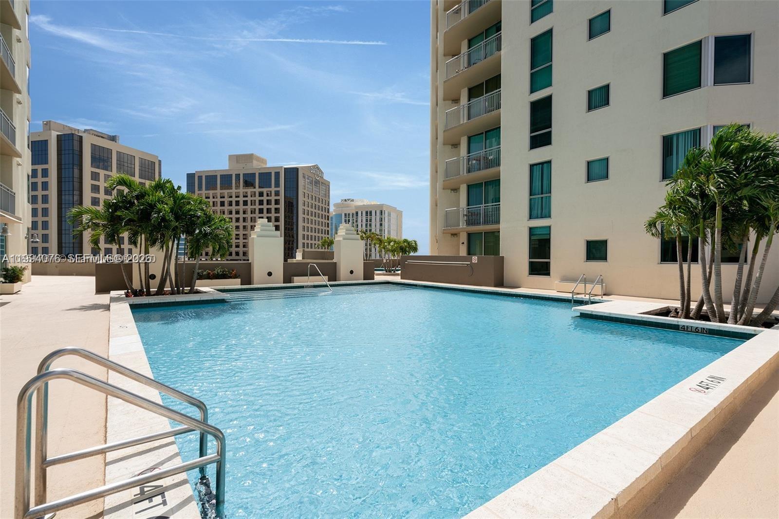 An outdoor swimming pool is situated between two tall residential buildings under a blue sky — 9066 SW 73rd Ct # 1202, Miami FL 33156, MIAMI, FL