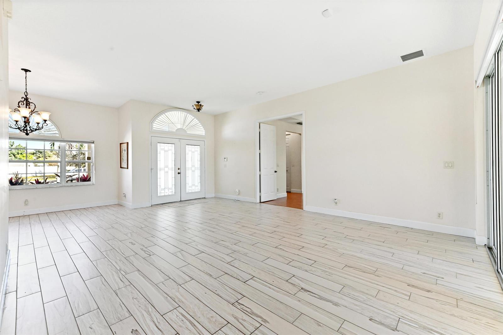 A bright, empty living room features light wood-look tile flooring, white walls, and double entry doors — 1791 Pierce Drive, FL 33460, FL