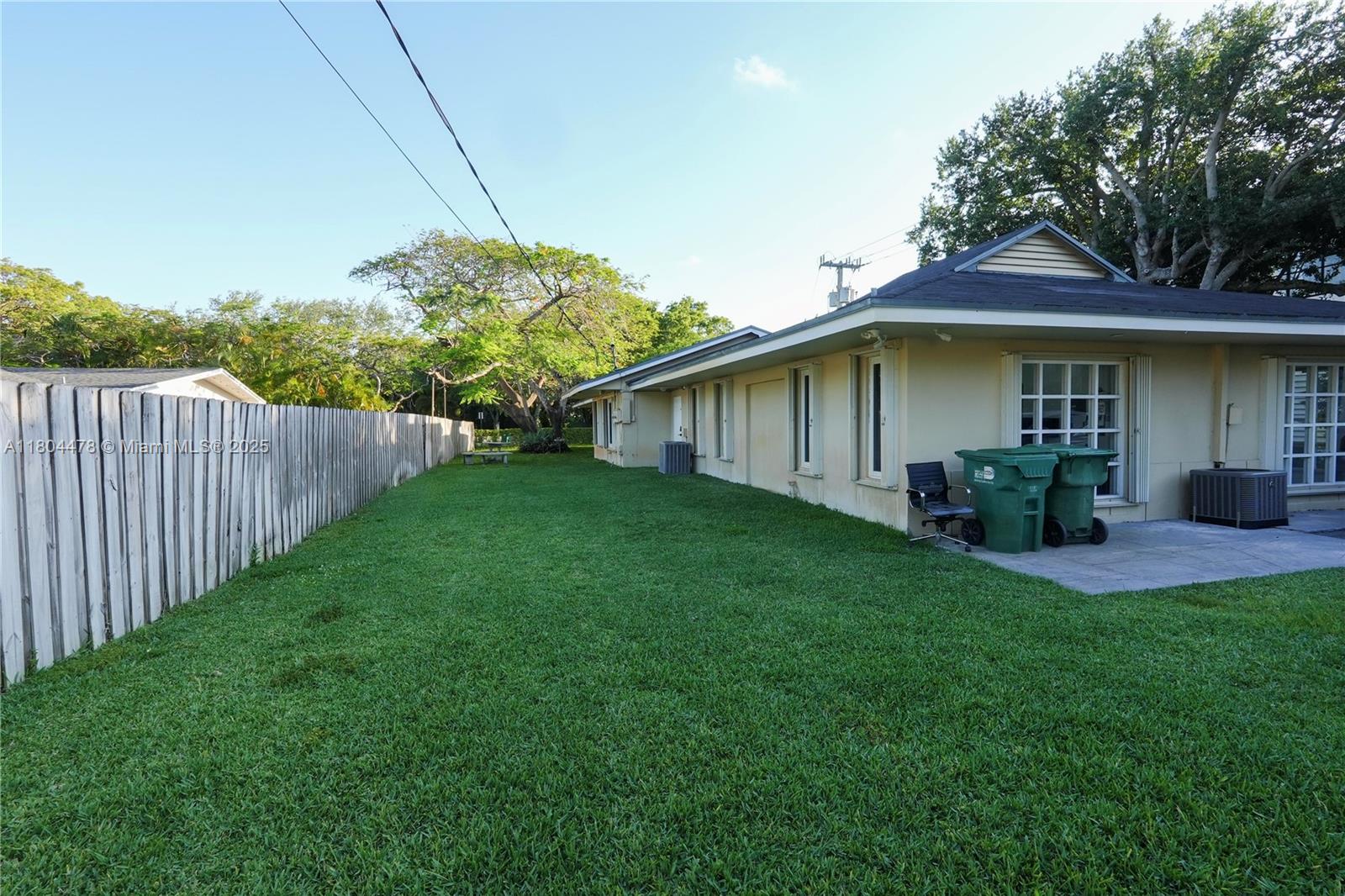 A side yard with a green lawn, a wooden fence, and the exterior of a house — 8125 SW 120th St, Pinecrest FL 33156, PINECREST, FL