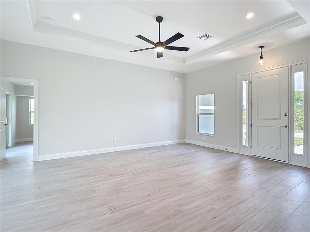 A bright, empty living room features light wood-look tile flooring, a tray ceiling, and a ceiling fan — 13503 AINSWORTH LANE, PORT CHARLOTTE, FL