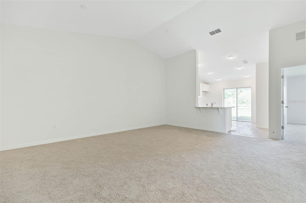 An empty living room with beige carpet and a vaulted ceiling opens to a kitchen — 20 BUTTONWOOD LANE, PALM COAST, FL