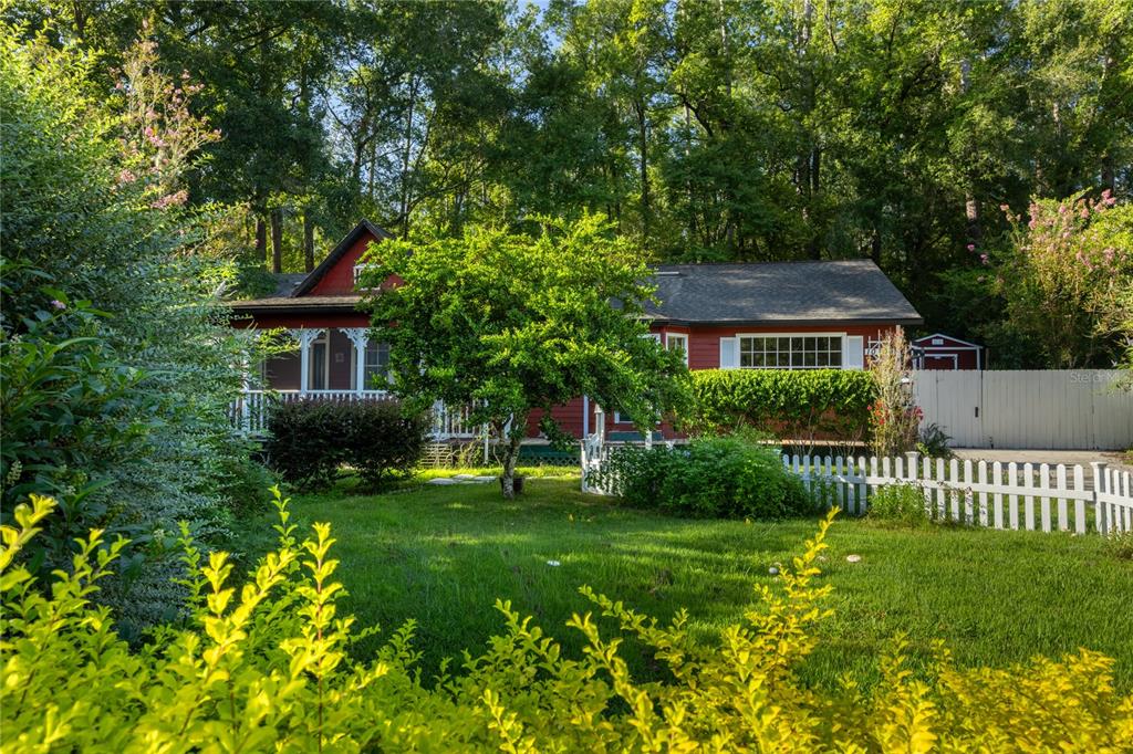 A red house with a white picket fence sits behind a green lawn and trees — 10709 NW 31ST PLACE, GAINESVILLE, FL