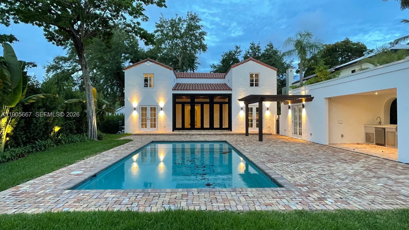 A swimming pool sits in front of a white house with a covered outdoor kitchen — 4100 Monserrate St, Coral Gables FL 33146, CORAL GABLES, FL