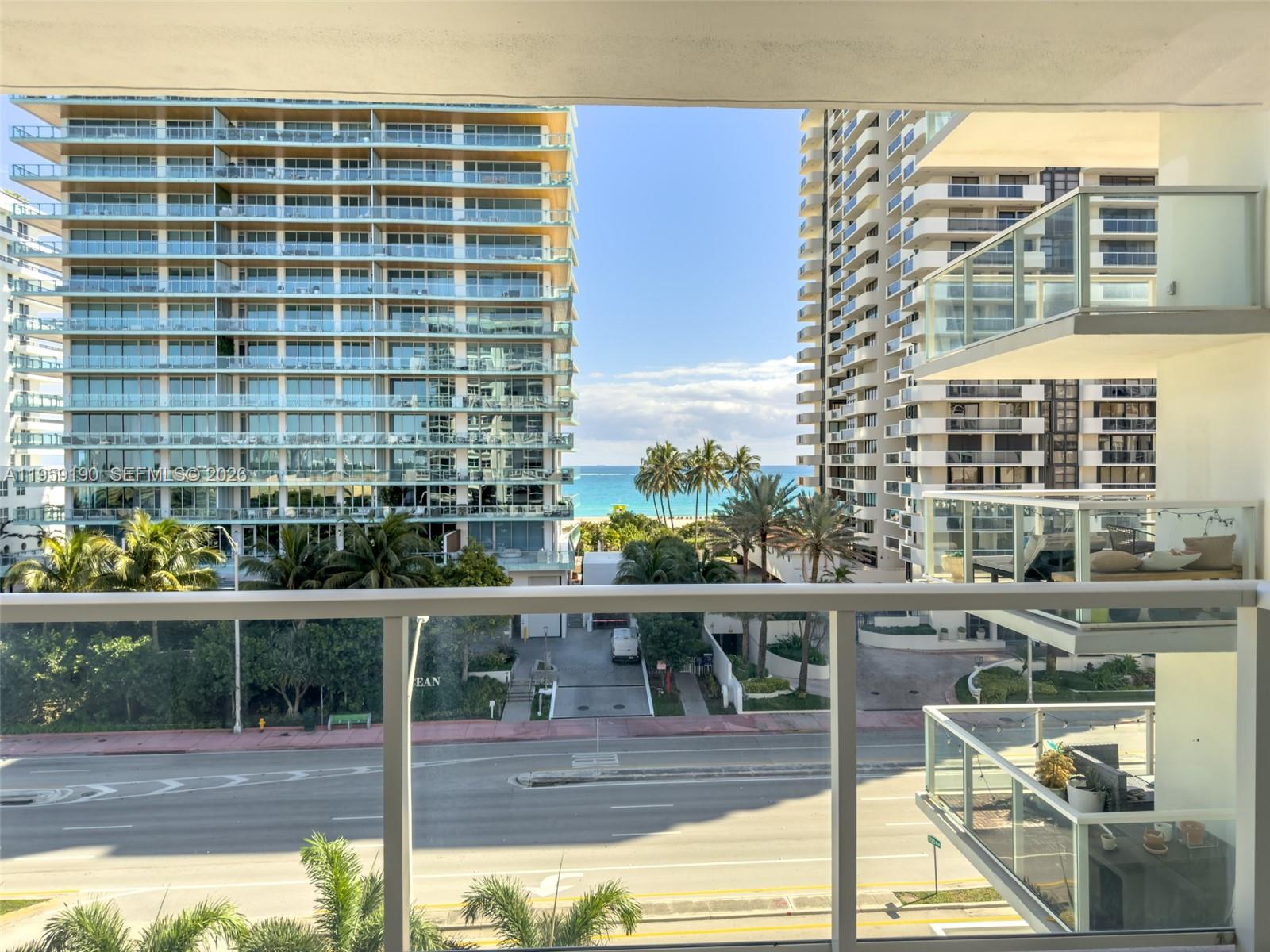 A balcony view overlooks a street, palm trees, the ocean, and two large apartment buildings — 5750 Collins Ave # 6J, Miami Beach FL 33140, MIAMI BEACH, FL