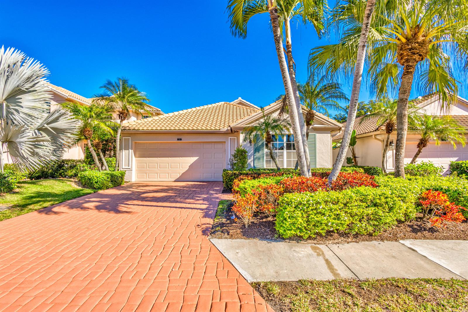 A single-story house with a tan tiled roof, beige stucco walls, and a brick driveway — 919 Augusta Pointe Drive, Palm Beach Gardens FL 33418, PALM BEACH GARDENS, FL