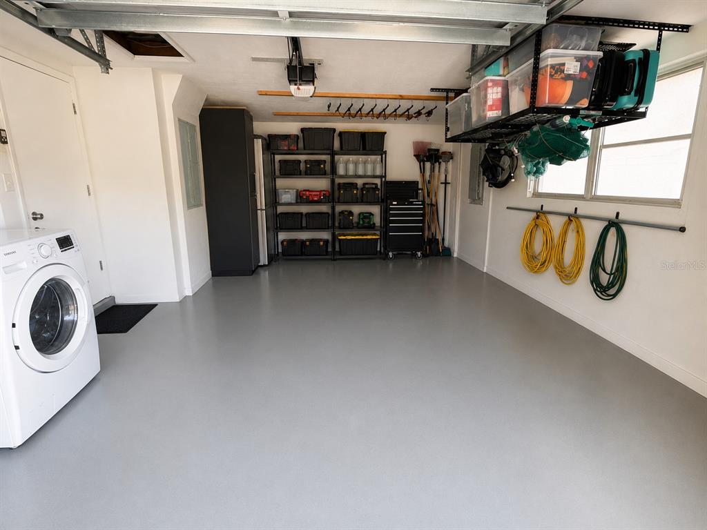 A garage with a gray epoxy floor, storage shelving, a washing machine, and wall-mounted hoses — 10124 HOLLY DRIVE, PORT RICHEY, FL