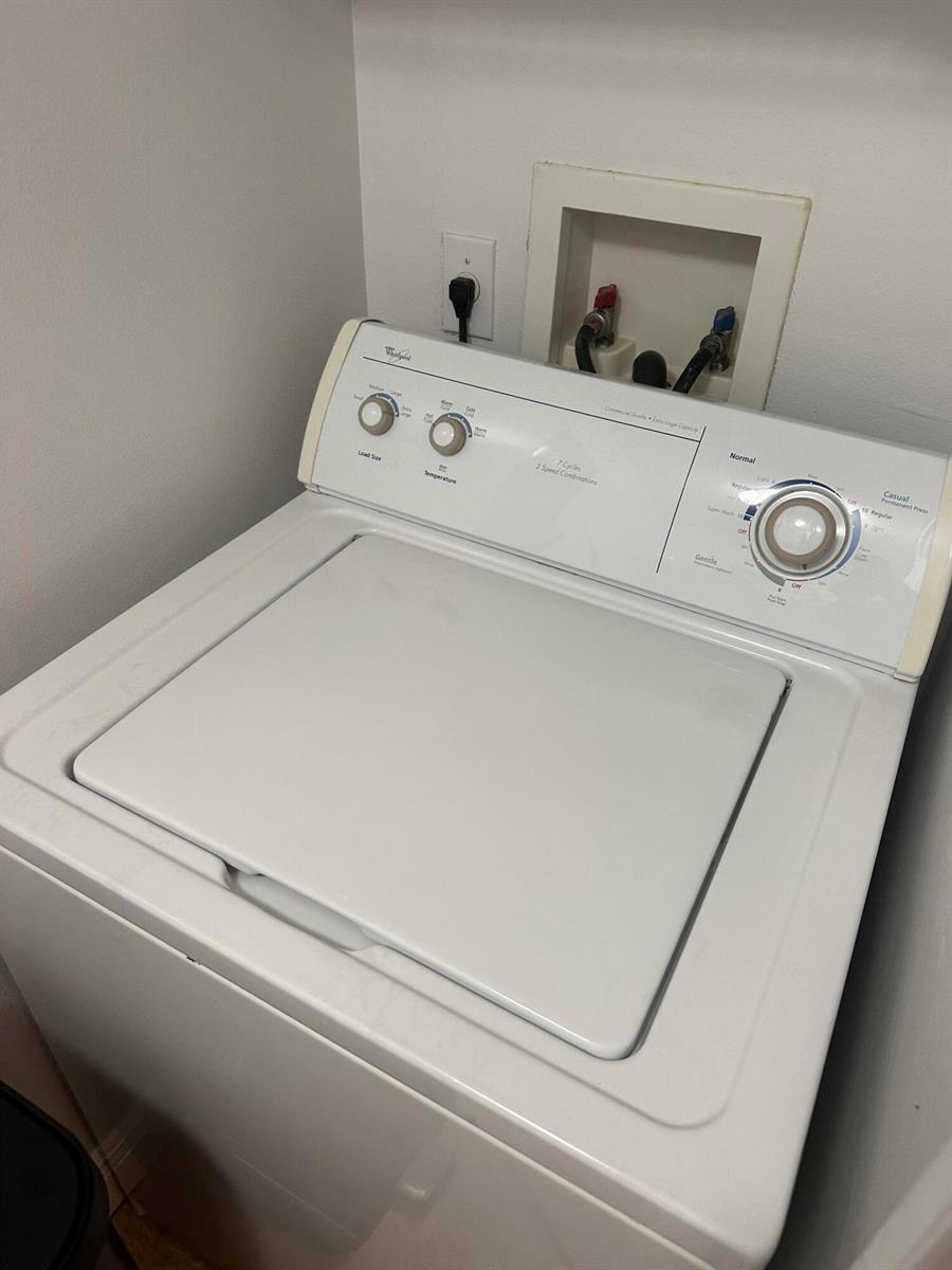 A white top-loading Whirlpool washing machine sits in a laundry area against a white wall — 400 Scotia Drive # 103, Hypoluxo FL 33462, HYPOLUXO, FL