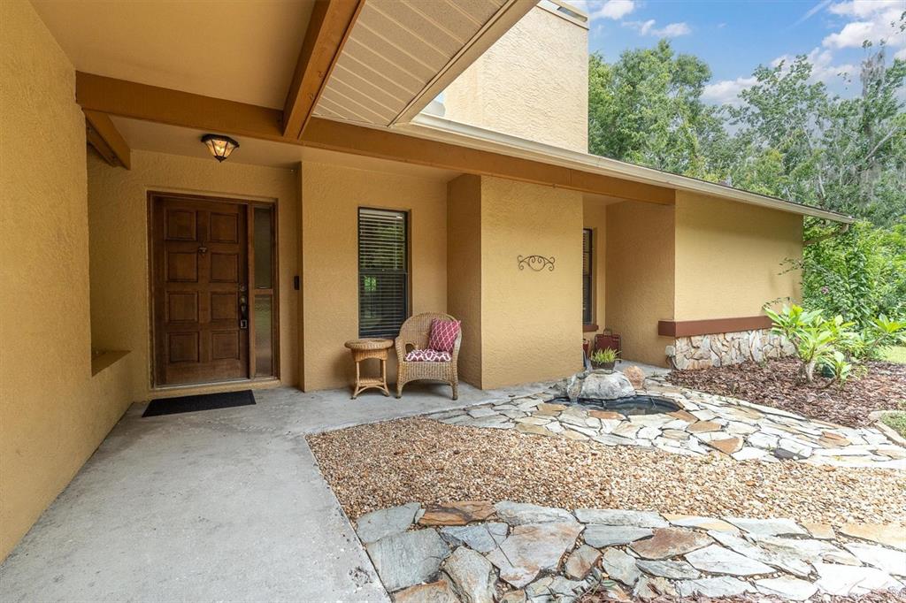 A covered entryway with a wooden door, wicker chair, and a small stone water feature — 6812 RIDGE TOP DRIVE, NEW PORT RICHEY, FL