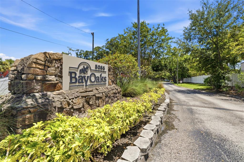 A stone monument sign for BayOaks stands beside a paved road lined with trees — 5265 E BAY DRIVE UNIT 114, CLEARWATER, FL