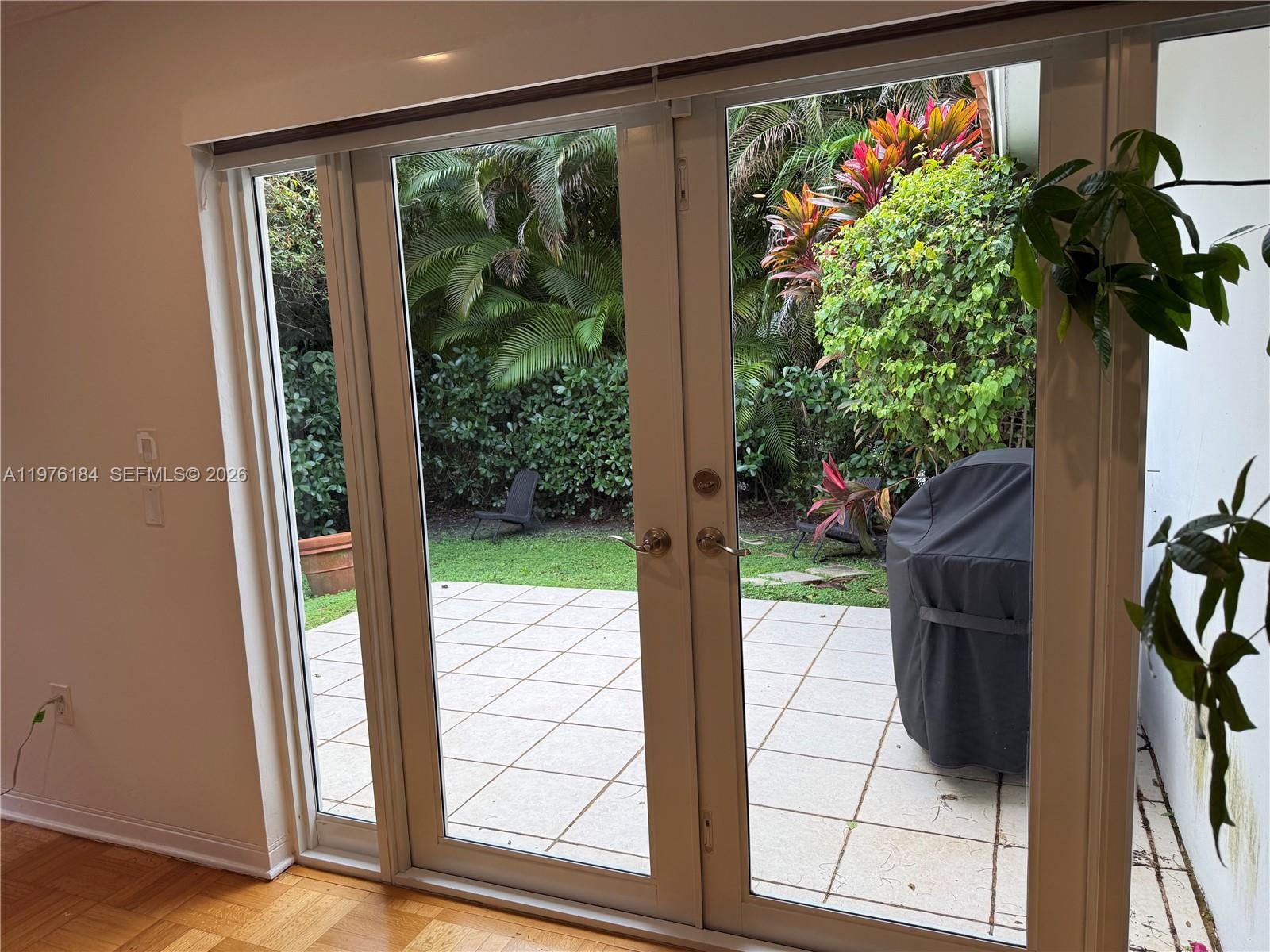 A view through glass double doors shows a tiled patio, a grill, and lush landscaping — 1428 Ortega Ave, Coral Gables FL 33134, CORAL GABLES, FL