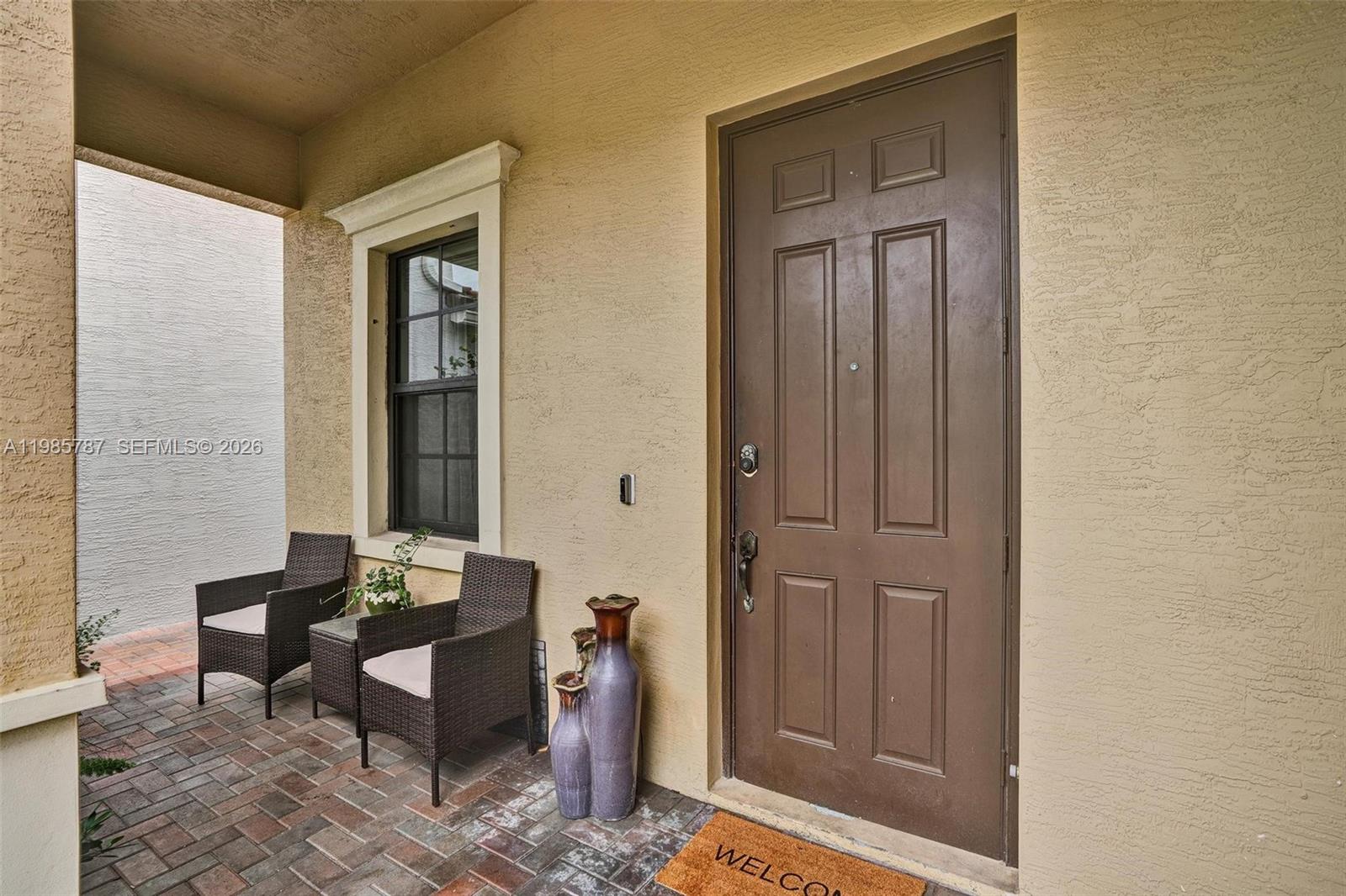 A covered entryway features a brown door, a window, two wicker chairs, and a vase — 760 NE 193rd Ter, Miami FL 33179, MIAMI, FL