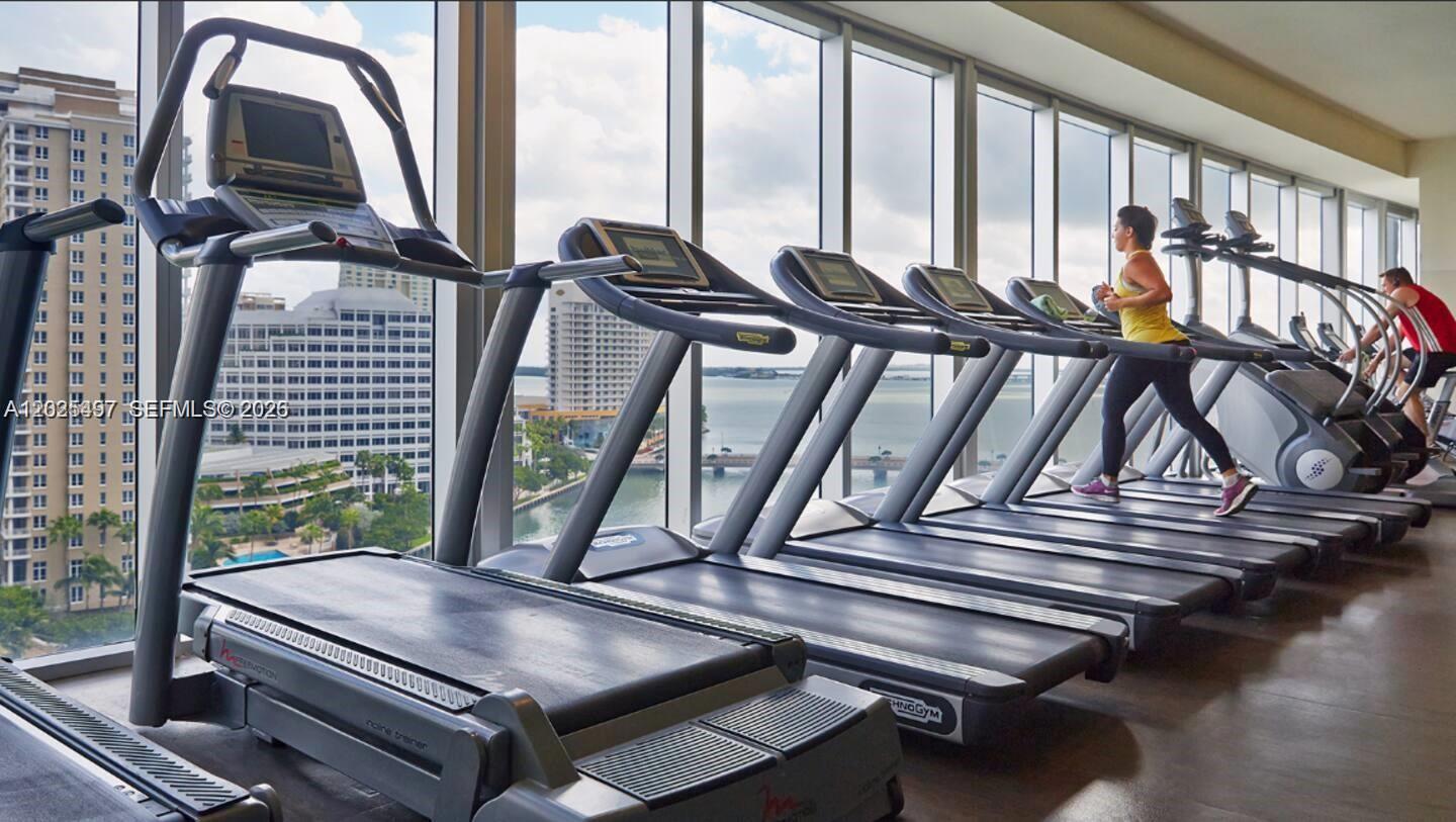 A gym with a row of treadmills facing floor-to-ceiling windows overlooking a bay and city — 485 Brickell Ave # 2006, Miami FL 33131, MIAMI, FL
