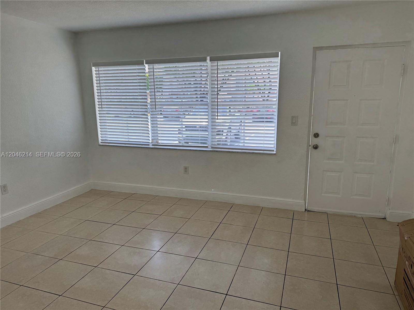 An empty room features white walls, tiled flooring, a large window with blinds, and a door — 8695 NW 6th Ln # 105, Miami FL 33126, MIAMI, FL