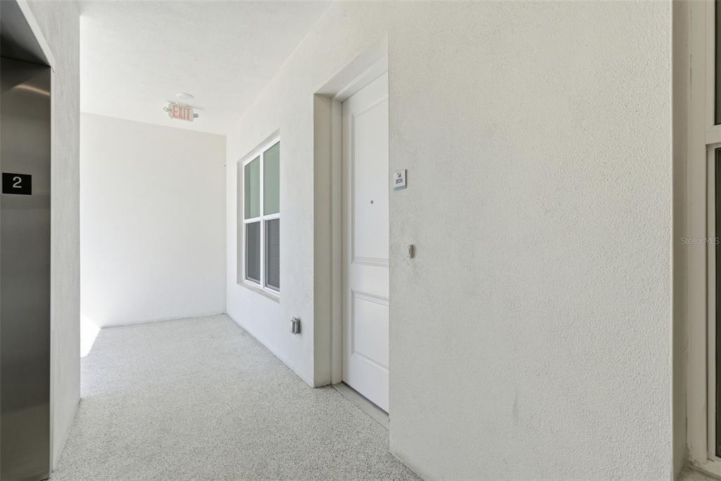 A white apartment hallway features a metal elevator door, a window, and a unit door — 400 150TH AVENUE UNIT 205, MADEIRA BEACH, FL