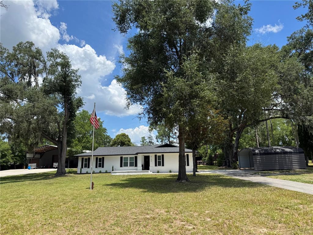 A white single-story house with black shutters and a metal carport sits on a lawn — 6133 SE 123RD LANE, BELLEVIEW, FL