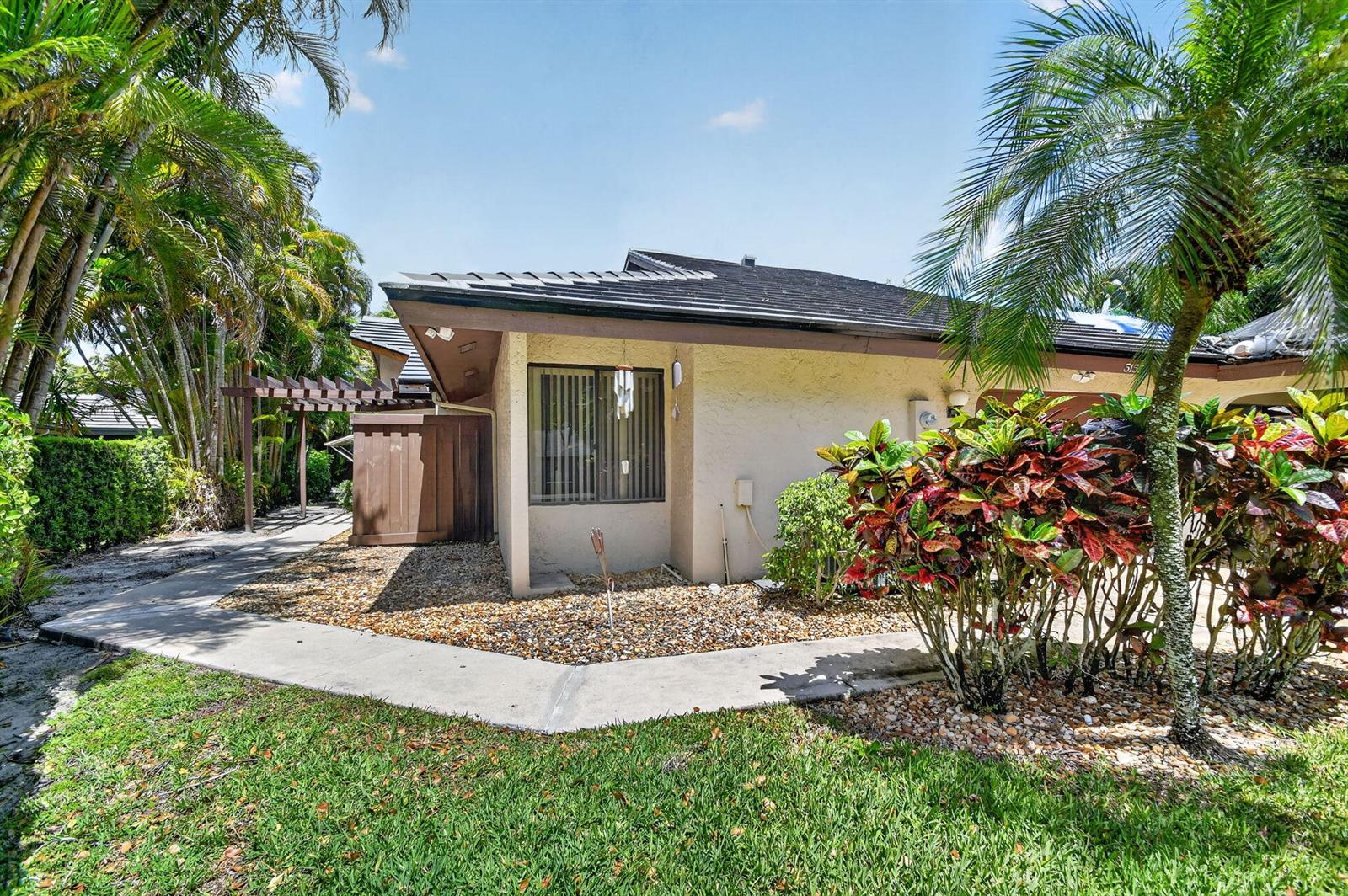 A single-story house exterior with a tan stucco wall, dark roof, and tropical landscaping — 5155 Pine Drive, Boynton Beach FL 33437, BOYNTON BEACH, FL