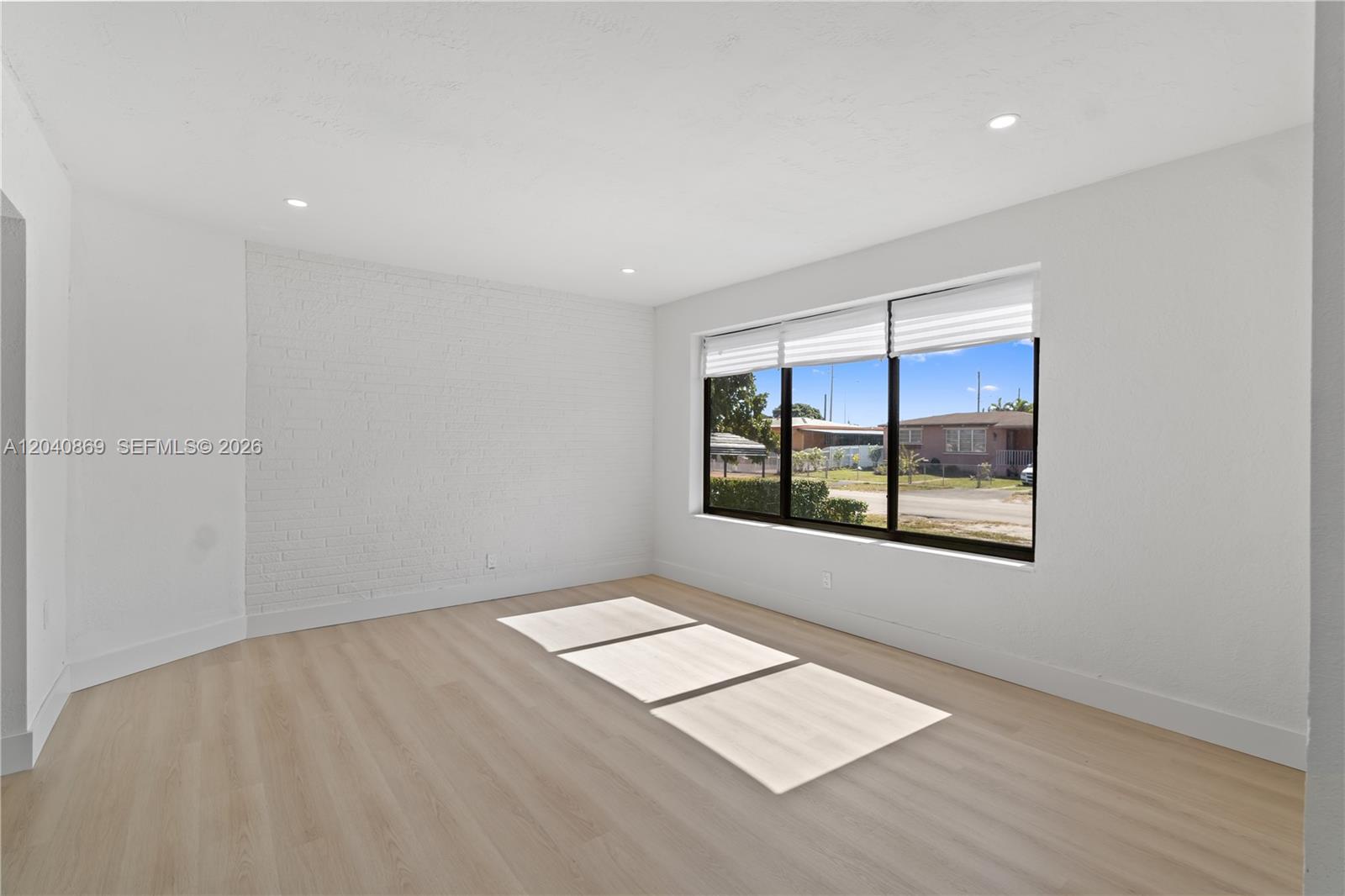 An empty room features light wood flooring, white walls, a brick accent wall, and large windows — 31 W 64th St, Hialeah FL 33012, HIALEAH, FL