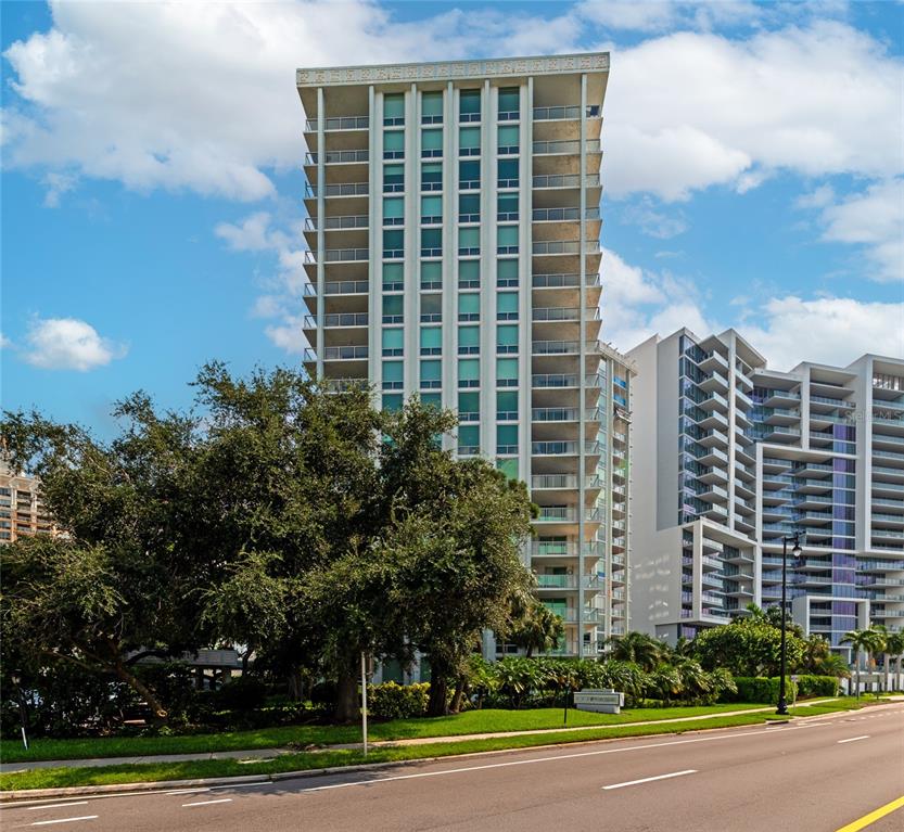 A tall, multi-story residential building stands behind a large tree and a paved road — 1111 N GULFSTREAM AVE #1A AVENUE UNIT 1A, SARASOTA, FL