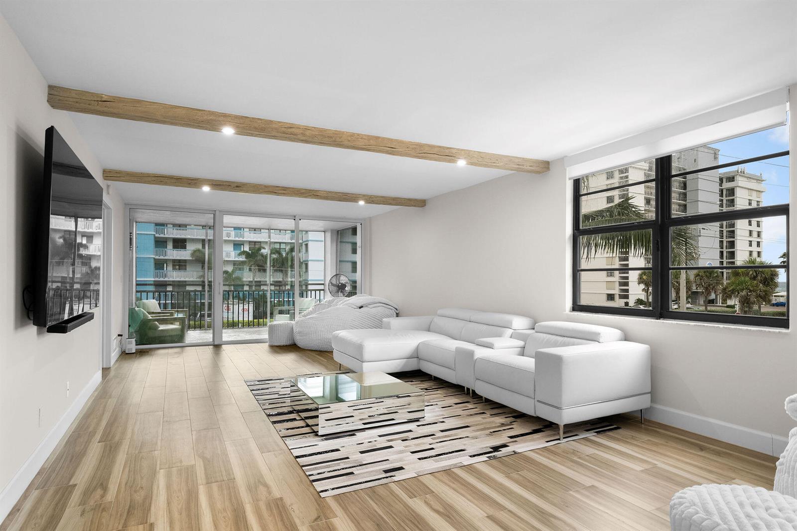 A living room features white leather furniture, wood-look flooring, exposed ceiling beams, and balcony access — 911 Ocean Drive # 201, Juno Beach FL 33408, JUNO BEACH, FL