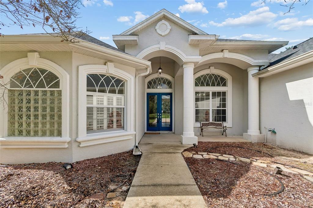 The exterior of a light-colored house features a covered entryway with columns and blue double doors — 35841 MIDDEN PATH, ZEPHYRHILLS, FL