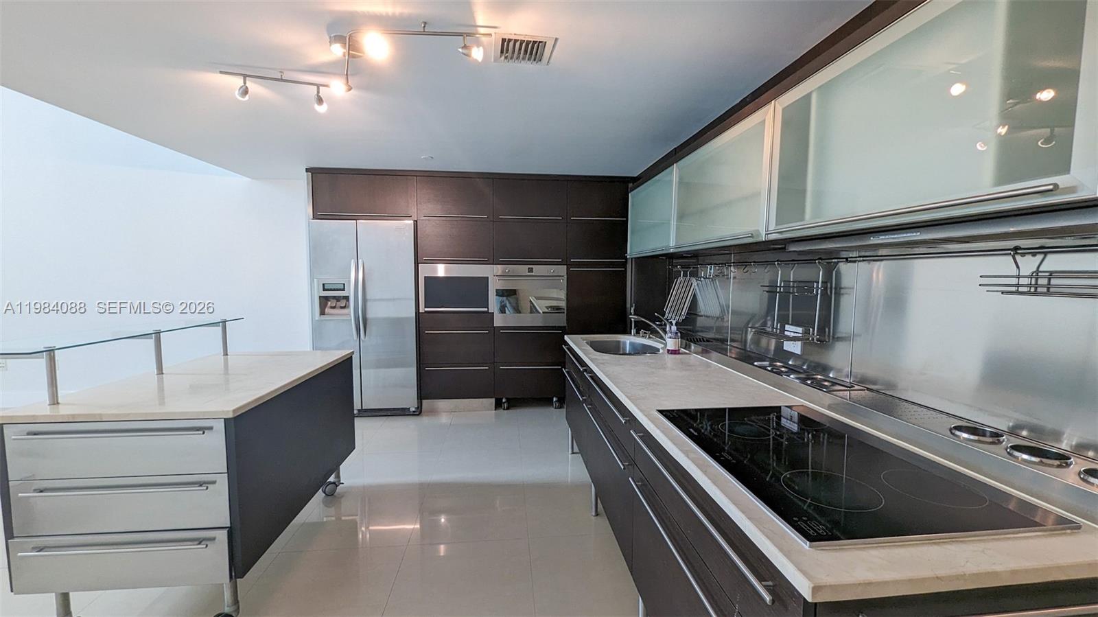 A modern kitchen features dark cabinetry, stainless steel appliances, and a mobile island — 6000 Collins Ave # 111, Miami Beach FL 33140, MIAMI BEACH, FL