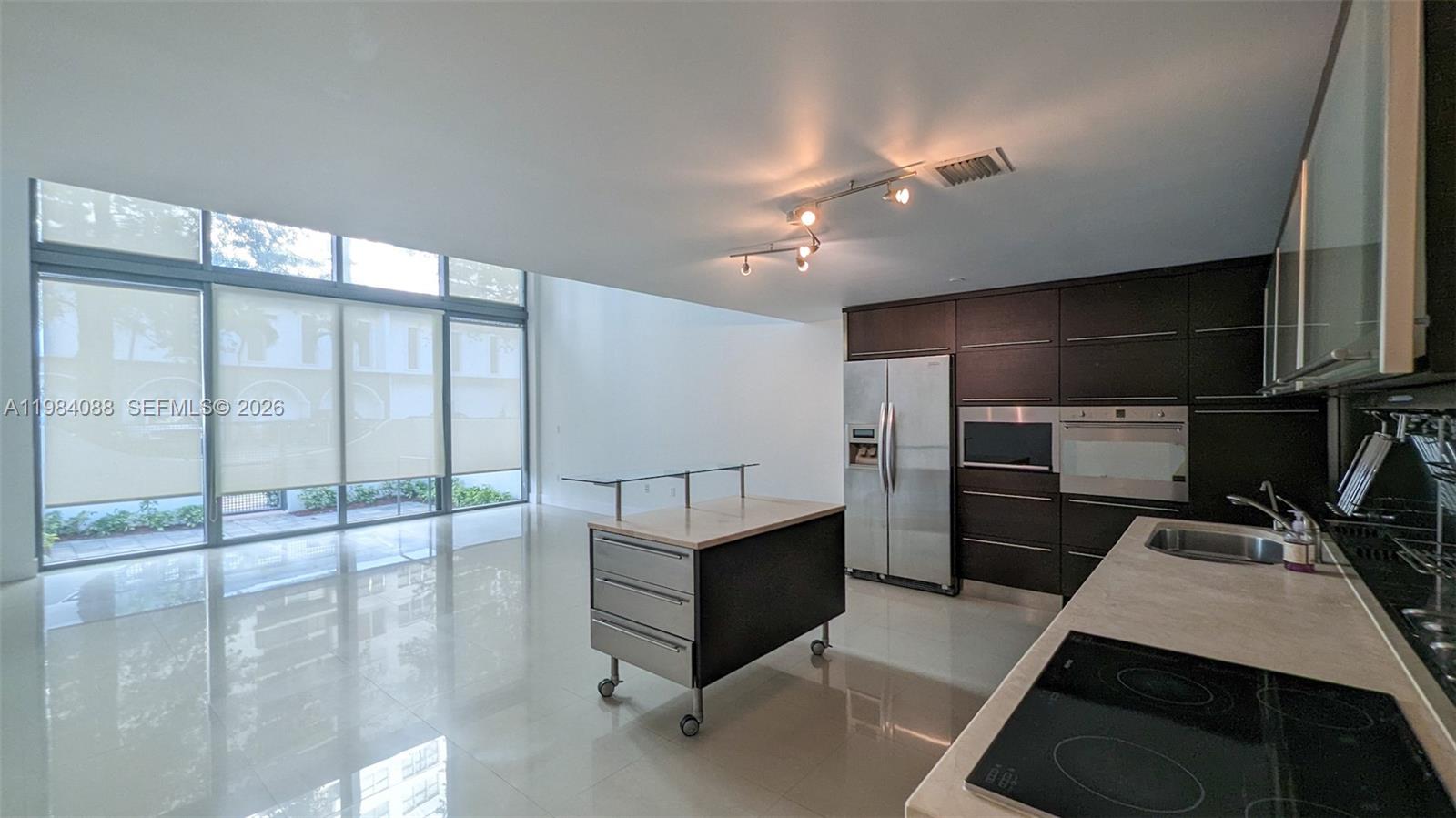 A modern kitchen with dark cabinetry and a mobile island overlooks a bright living area — 6000 Collins Ave # 111, Miami Beach FL 33140, MIAMI BEACH, FL