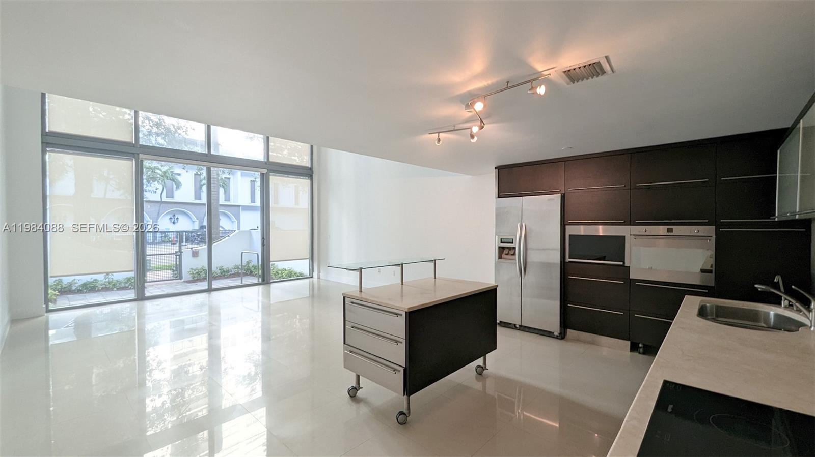 A modern kitchen with dark cabinetry and a rolling island opens to a living area — 6000 Collins Ave # 111, Miami Beach FL 33140, MIAMI BEACH, FL