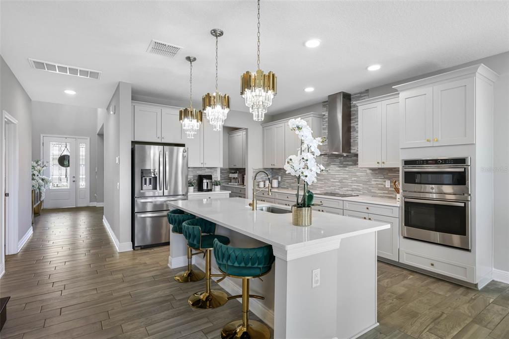 A modern kitchen features white cabinetry, a large island with teal stools, and stainless appliances — 38144 COBBLE CREEK BOULEVARD, ZEPHYRHILLS, FL