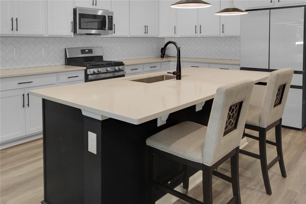 A modern kitchen features white cabinets, a dark island with bar stools, and stainless appliances — 5208 MEADOW SONG DRIVE, OKAHUMPKA, FL