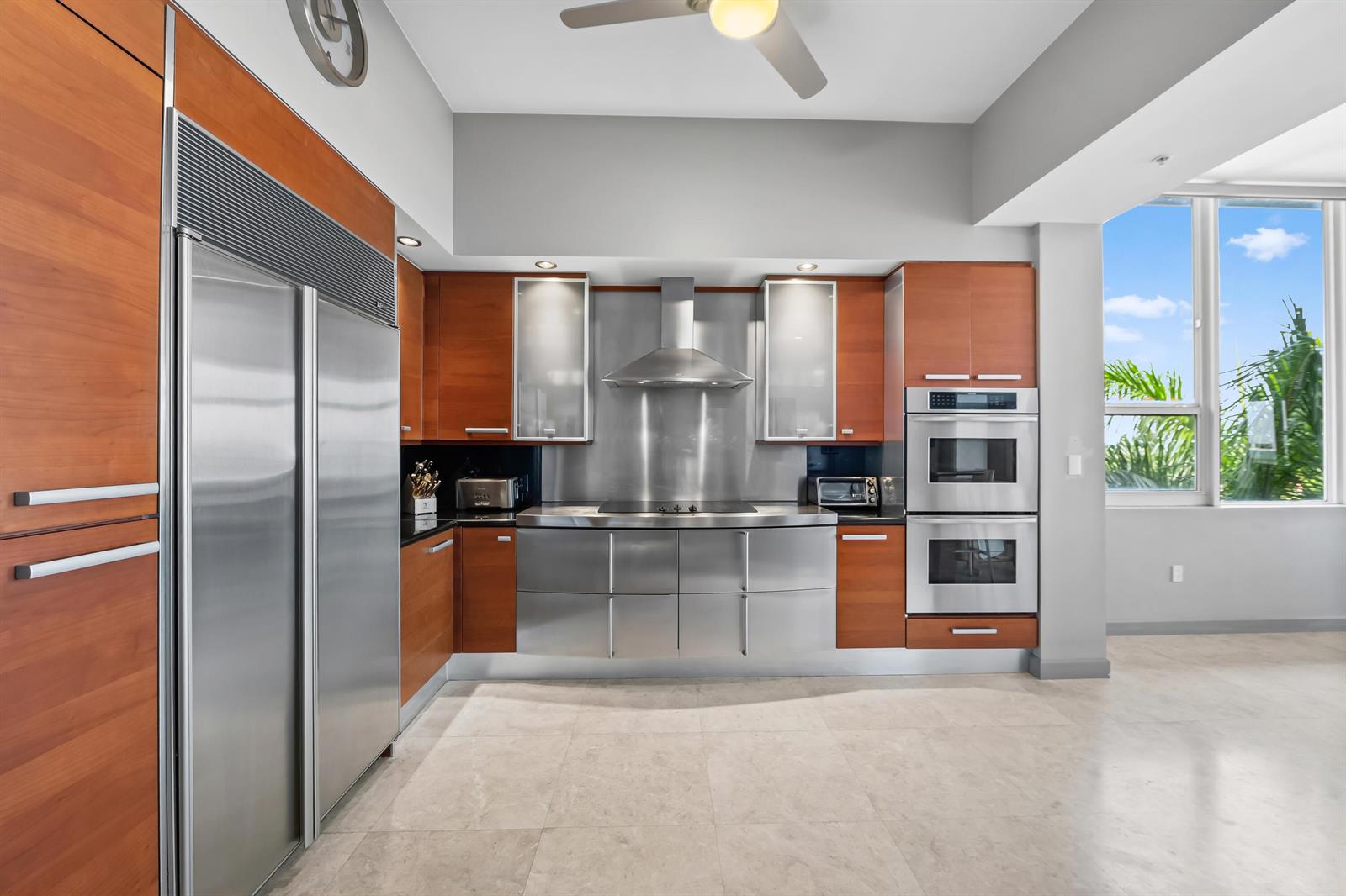 A modern kitchen features wood cabinetry, stainless steel appliances, and a large window with greenery — 701 SE 21st Avenue # 501, Deerfield Beach FL 33441, DEERFIELD BEACH, FL