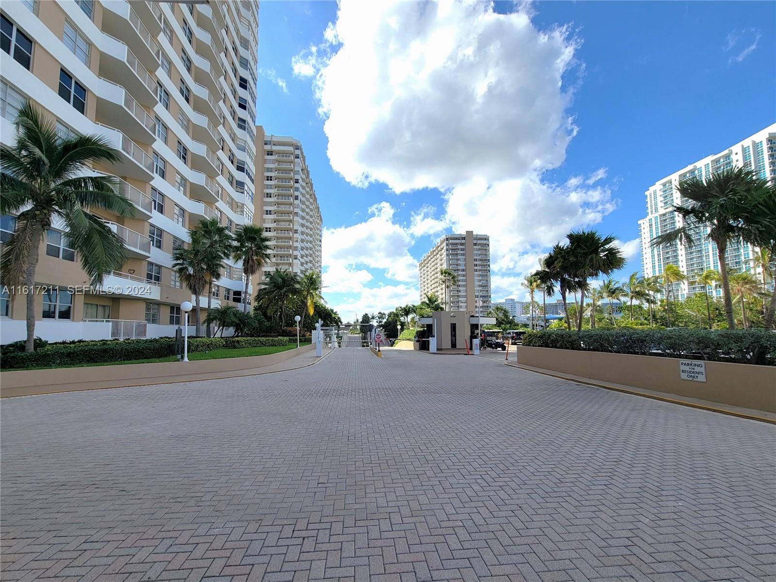 A paved driveway leads toward a gated entrance between two tall residential apartment buildings — 1980 S Ocean Dr # 2C, Hallandale Beach FL 33009, HALLANDALE BEACH, FL