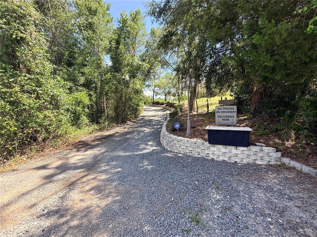 A gravel driveway leads uphill past a stone retaining wall and a delivery sign — 25962 WHIPPERWILL STREET, BROOKSVILLE, FL
