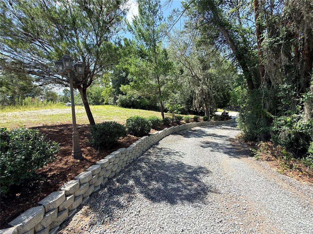 A gravel driveway curves past a retaining wall and a lamp post toward a wooded area — 25962 WHIPPERWILL STREET, BROOKSVILLE, FL
