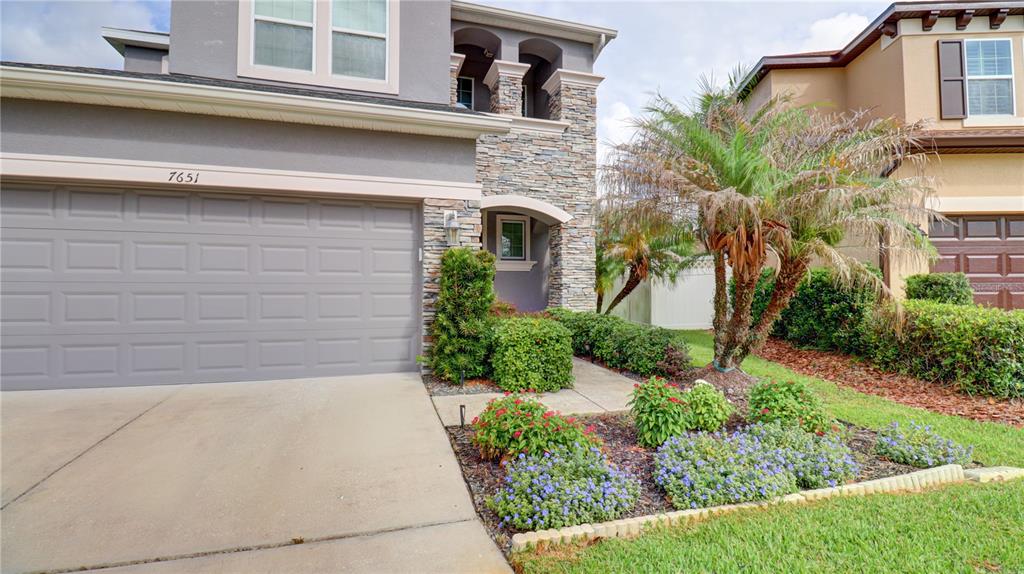 A two-story house exterior features a gray garage, stone accents, and a landscaped front yard — 7651 WINDCHASE WAY, WESLEY CHAPEL, FL