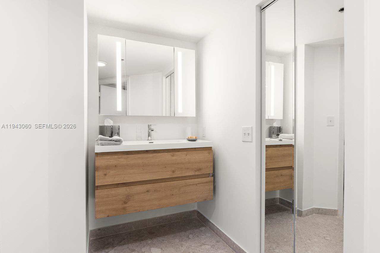 A modern bathroom features a floating wood vanity, a white countertop, and a mirrored cabinet — 6767 Collins Ave # 1905, Miami Beach FL 33141, MIAMI BEACH, FL