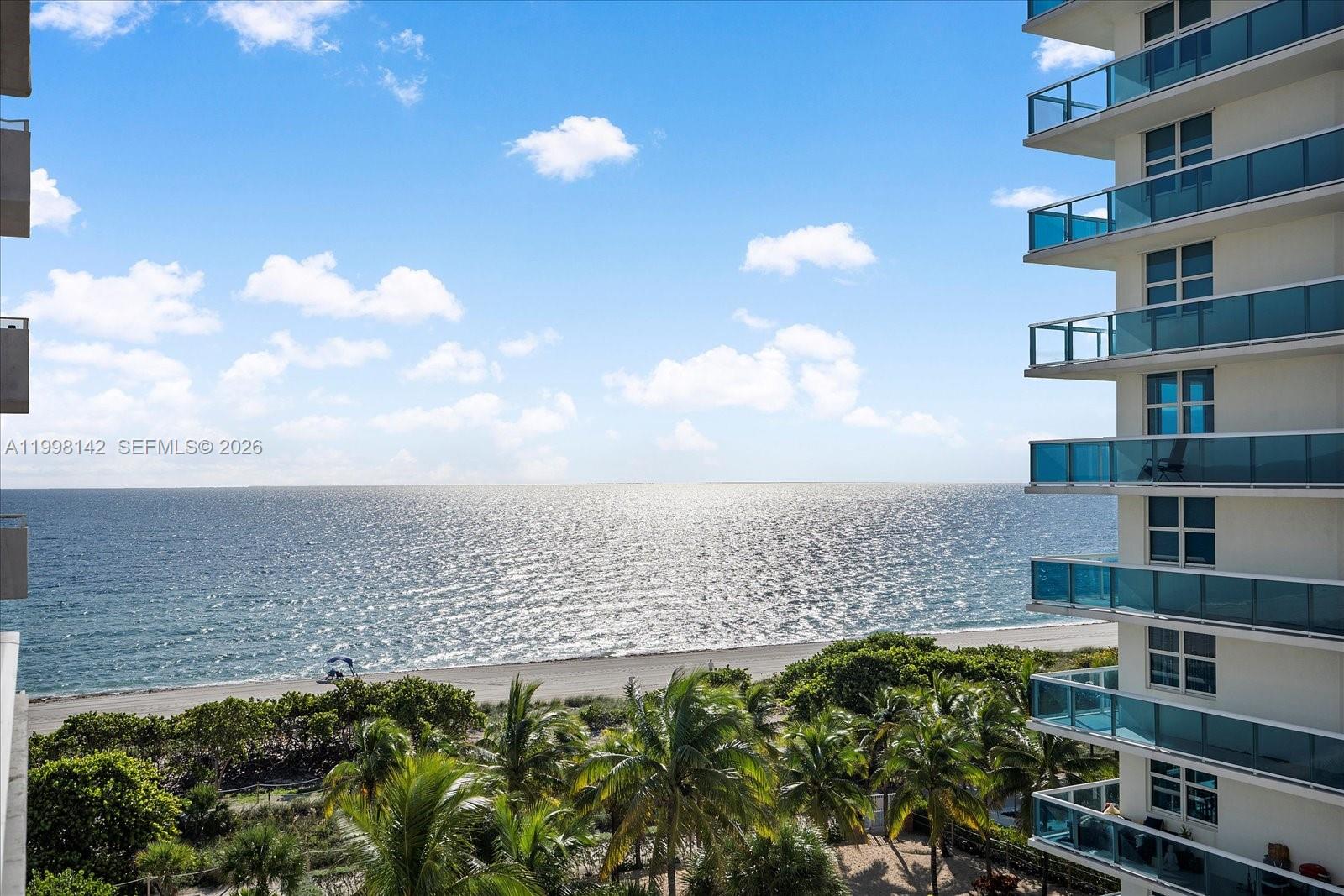 A high-rise building overlooks a sandy beach, palm trees, and the ocean under a blue sky — 9225 Collins Ave # 706, Surfside FL 33154, SURFSIDE, FL