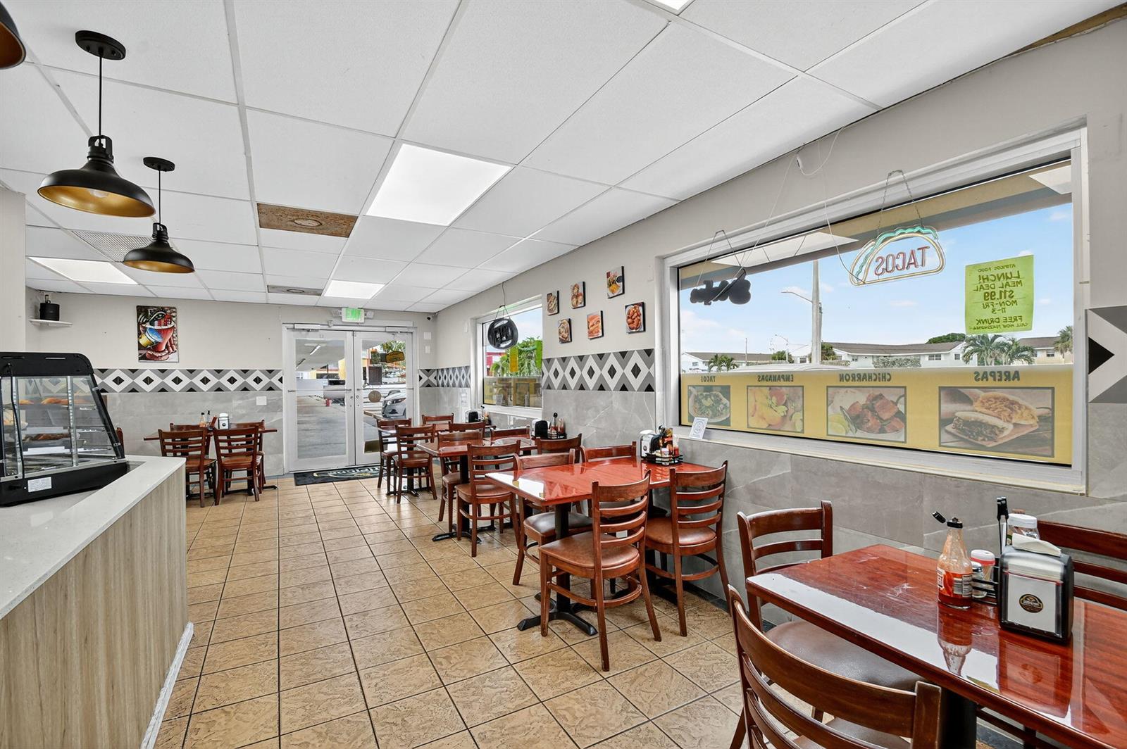 A brightly lit restaurant interior features tiled floors, wooden tables, and chairs near large windows — 1541 W Blue Heron Boulevard, Riviera Beach FL 33404, RIVIERA BEACH, FL
