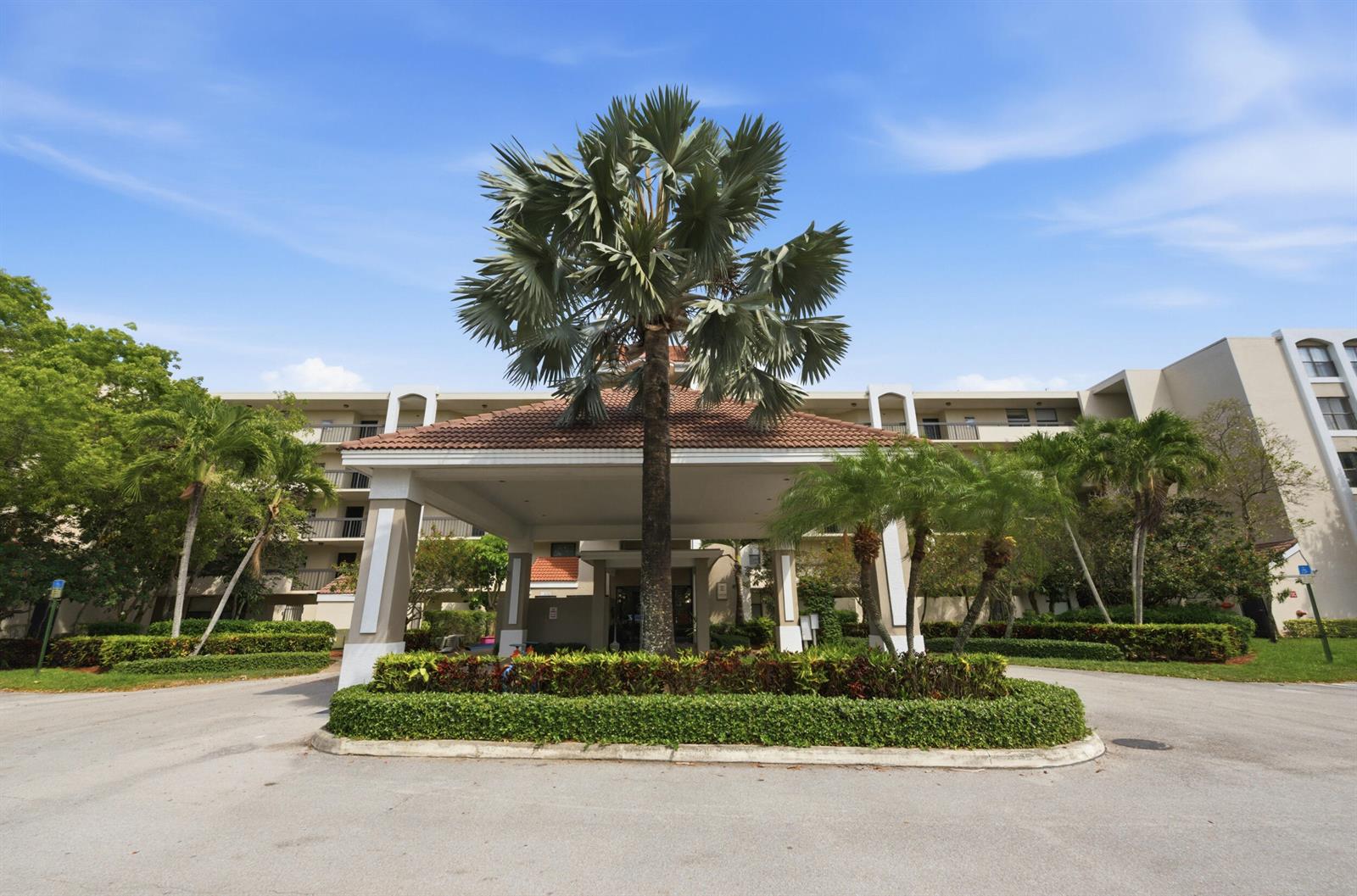 A palm tree stands in front of a building entrance with a covered driveway — 1915 Lavers Circle # E508, Delray Beach FL 33444, DELRAY BEACH, FL