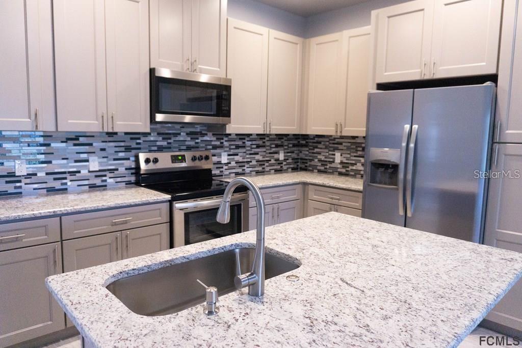 A kitchen features gray cabinets, granite countertops, a stainless steel island sink, and mosaic backsplash — 59 FANSHAWE LANE UNIT B, PALM COAST, FL