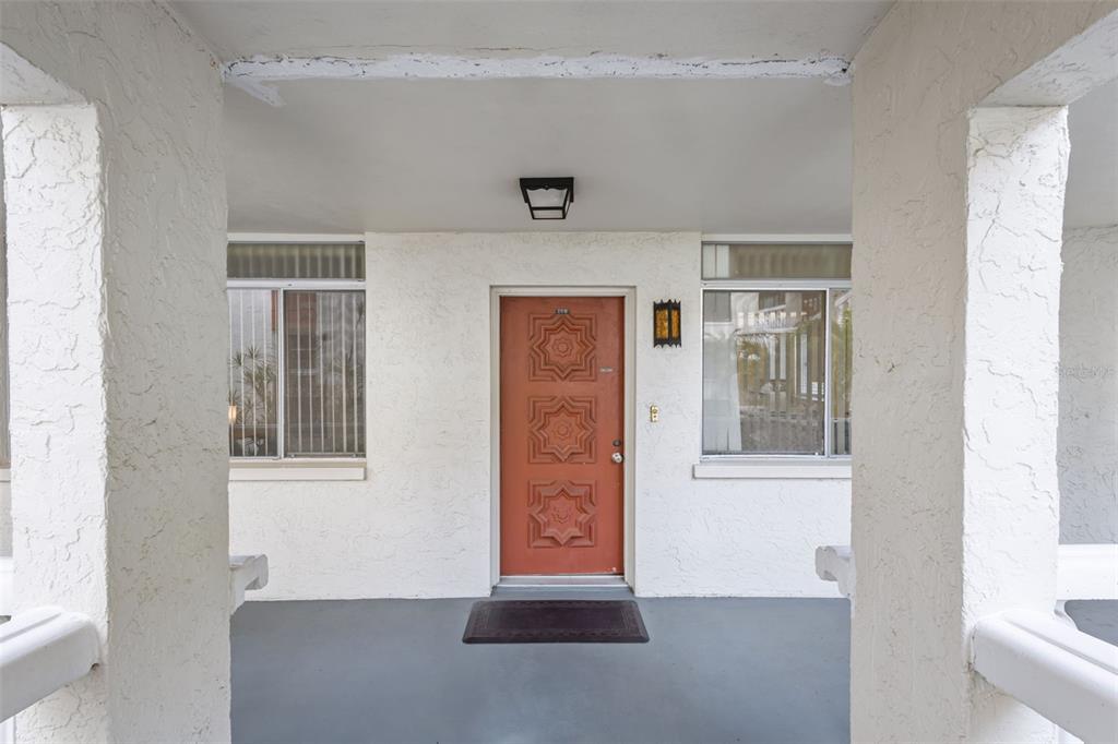 A covered porch features a textured white exterior wall, two windows, and a decorative orange door — 1009 PEARCE DRIVE UNIT 206, CLEARWATER, FL