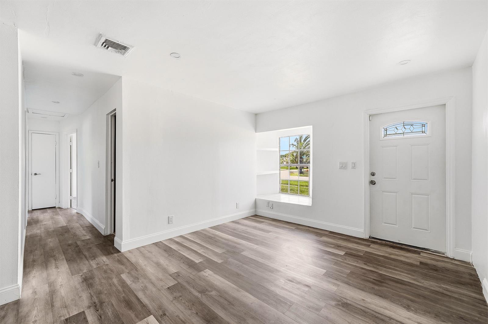 A bright, empty room features white walls, wood-look flooring, a window seat, and a front door — 1060 SW 5th Terrace, Deerfield Beach FL 33441, DEERFIELD BEACH, FL