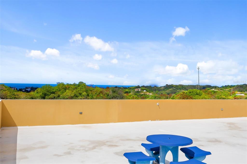 A rooftop terrace features a blue table and chairs overlooking a lush landscape and ocean — Km 15 H 7 PR 115 VILLA LA JOLLA, RINCON, PR