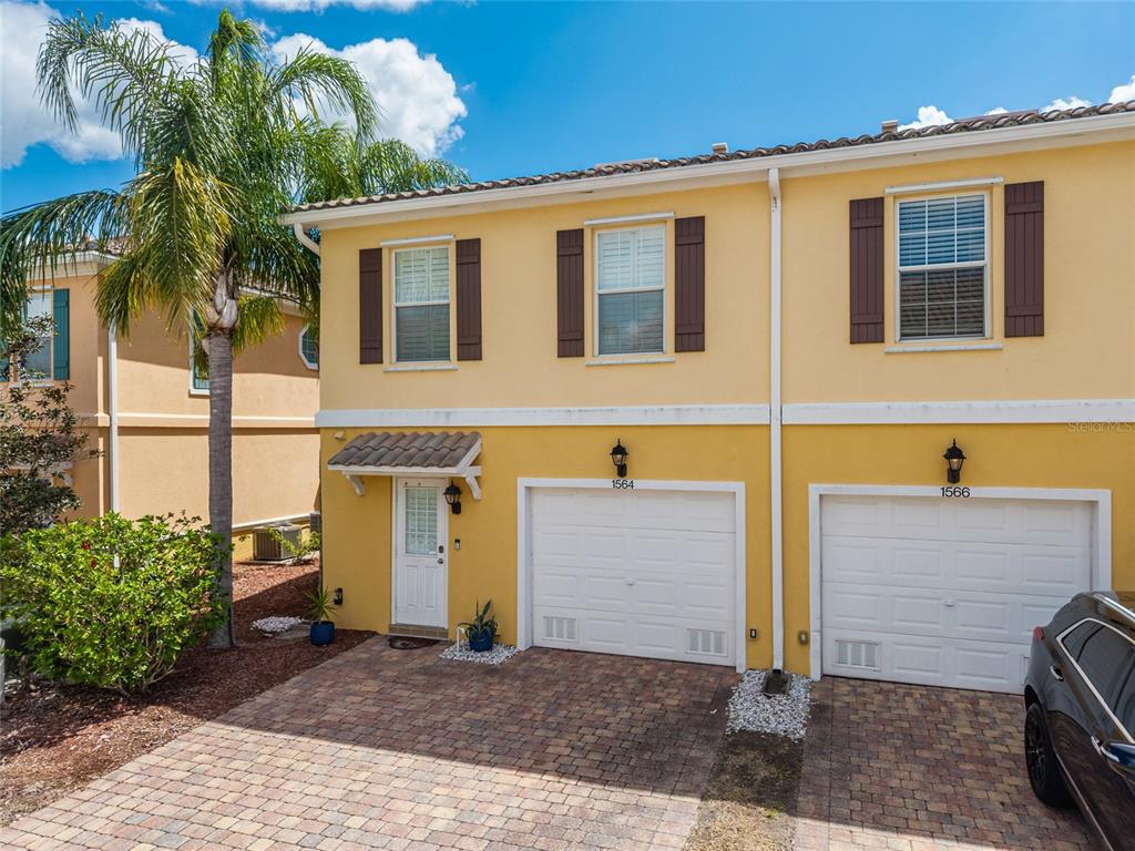 A two-story yellow townhouse with a brick driveway, white garage doors, and a palm tree — 1564 NAPOLI DRIVE W, SARASOTA, FL