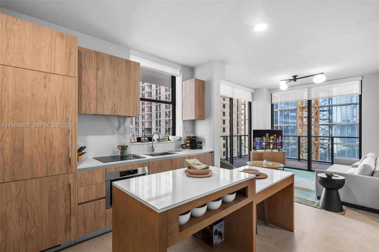 A modern kitchen with wood cabinetry and a white island opens into a living area — 11 NE 6th # 2207, Miami FL 33131, MIAMI, FL