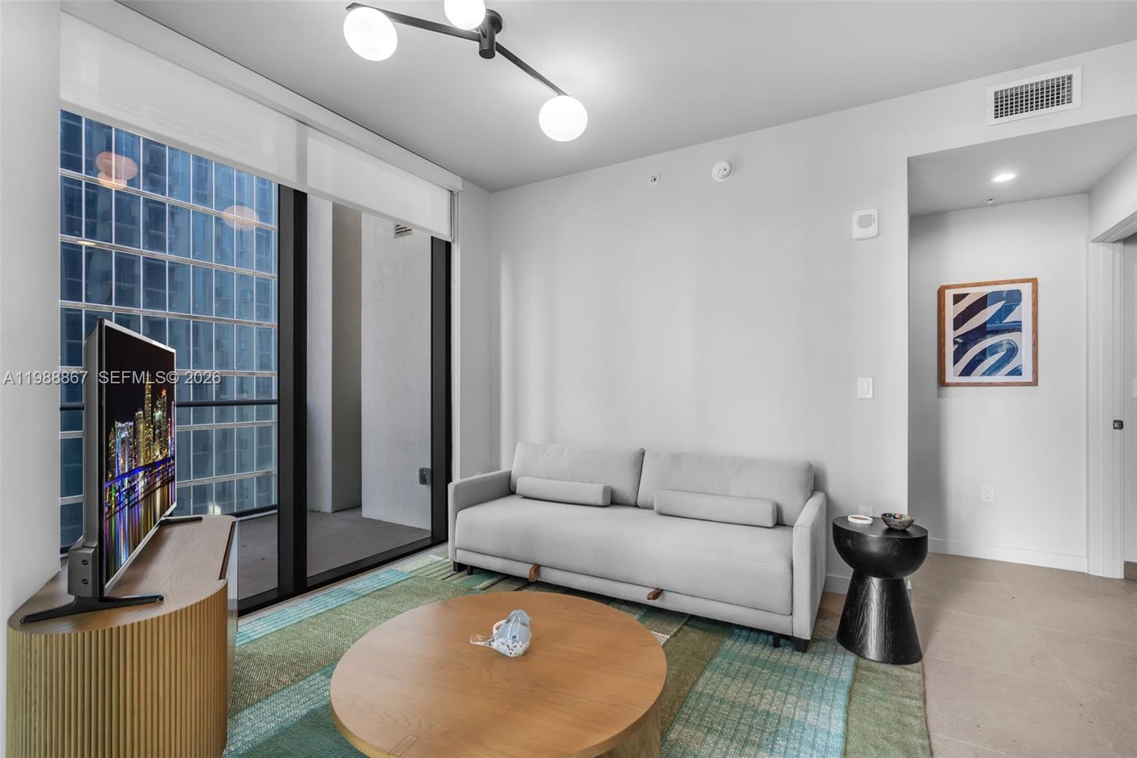 A modern living room features a light gray sofa, round coffee table, and floor-to-ceiling windows — 11 NE 6th # 2207, Miami FL 33131, MIAMI, FL