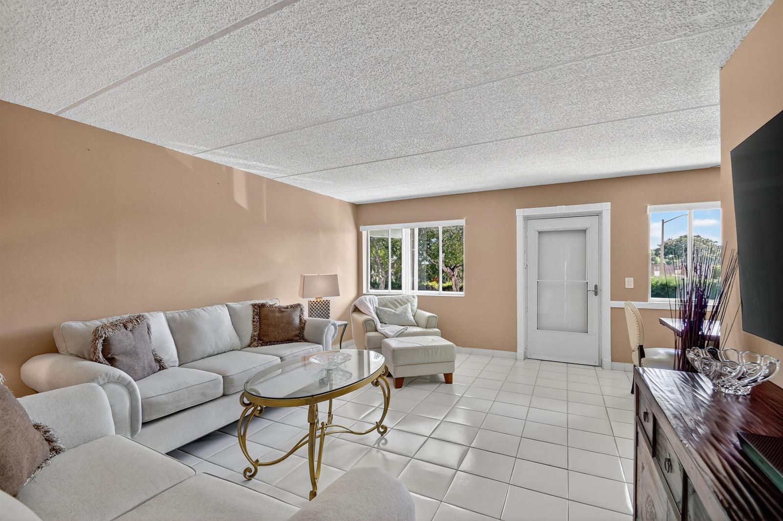 A living room features white tile flooring, a beige sofa, a glass coffee table, and windows — 3200 Springdale Boulevard # 217, Palm Springs FL 33461, PALM SPRINGS, FL