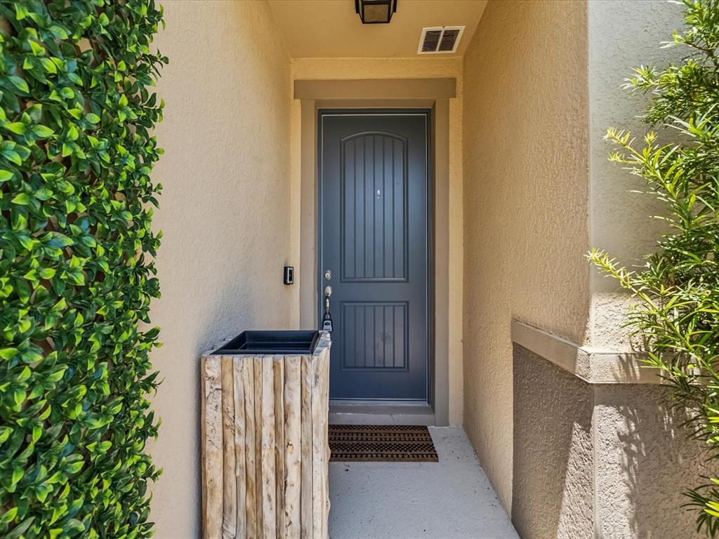 A dark gray front door is framed by beige stucco walls, a wooden planter, and greenery — 11525 SOUTHERN CREEK DRIVE, GIBSONTON, FL