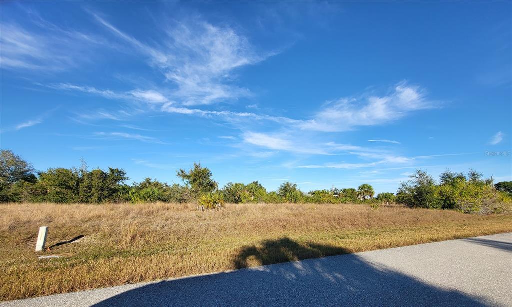 A vacant, grassy lot sits alongside a paved road under a clear blue sky — 13574 ALLAMANDA CIRCLE, PORT CHARLOTTE, FL